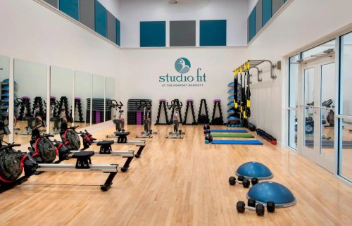 A bright fitness studio with exercise equipment, including weights, mats, and rowing machines, against a mirrored wall.