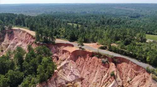 Aerial view of a rugged landscape with steep red cliffs, lush green trees, and a winding road along the edge.