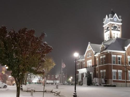 Snow-covered town square at night, featuring a historic clock tower and a tree with red leaves.