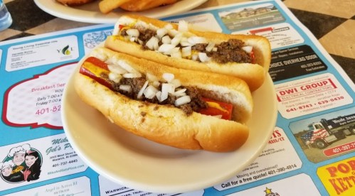 Two hot dogs topped with mustard, chopped onions, and a meat sauce, served on a plate over a colorful menu background.