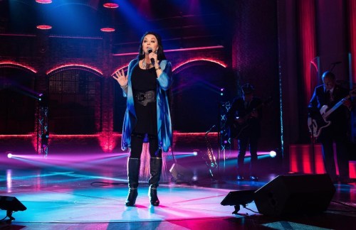 A singer performs on stage with colorful lighting, accompanied by a band in the background.