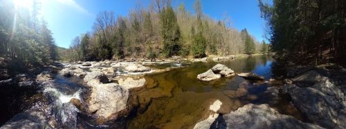 A serene river scene with clear water, rocky banks, and trees lining the shore under a bright blue sky.