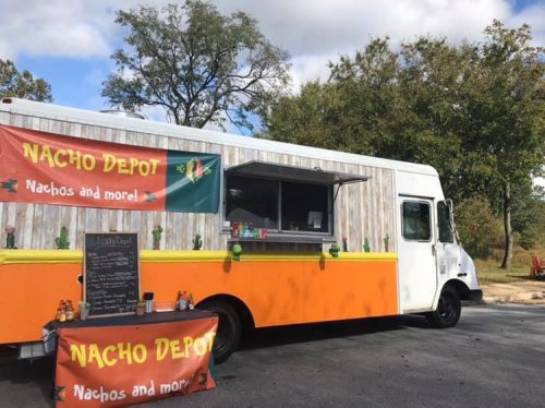 A colorful food truck named "Nacho Depot" with a menu board and outdoor seating under a blue sky.