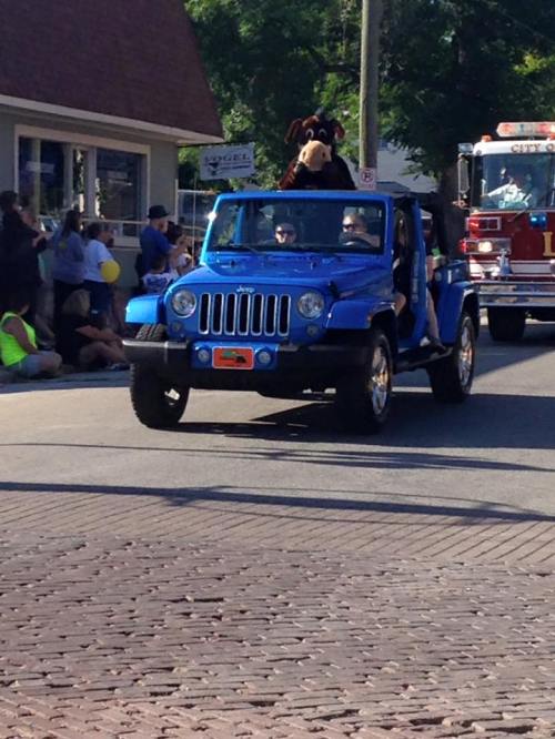 A blue jeep in a parade with a person in a mascot costume and onlookers enjoying the event.