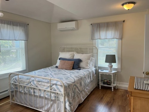Cozy bedroom with a white metal bed, patterned bedding, a nightstand, lamp, and natural light from two windows.