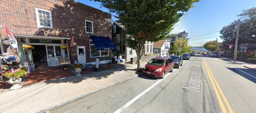 A quaint street scene featuring shops, parked cars, and trees, with a glimpse of water in the background.