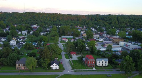 Aerial view of a small town with green trees, residential buildings, and a few commercial structures under a sunset sky.