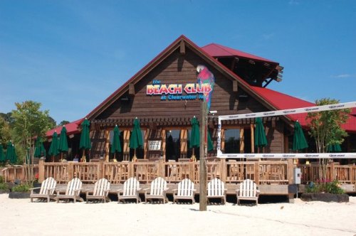 A beach club with a wooden exterior, green umbrellas, and white chairs on a sandy beach under a clear blue sky.
