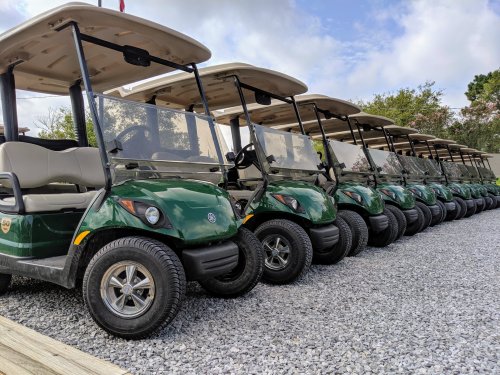 A row of green golf carts parked on a gravel surface under a cloudy sky.