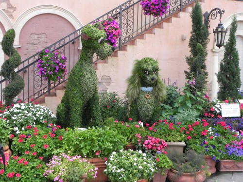Topiary sculptures of two dogs surrounded by colorful flowers and greenery, set against a charming staircase backdrop.