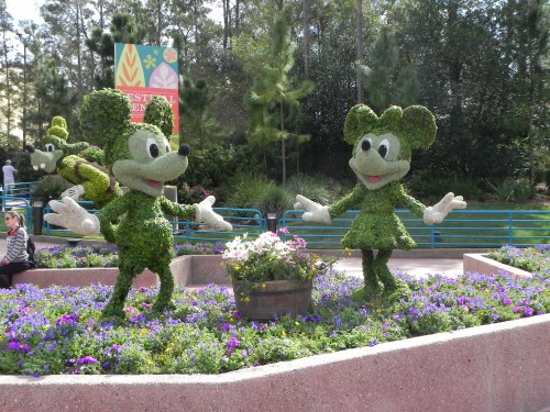 Topiary figures of Mickey and Minnie Mouse in a garden, surrounded by colorful flowers and greenery.