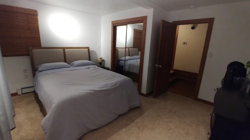 A cozy bedroom featuring a bed with gray bedding, a mirror, and a door leading to another room. Soft lighting enhances the space.