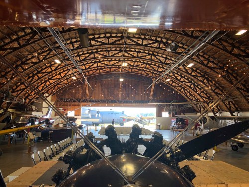 Interior of a large hangar with vintage aircraft and a wooden arched ceiling, viewed from the front of a plane.