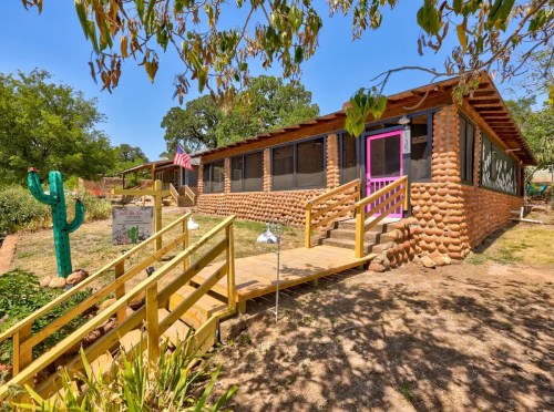 A rustic, adobe-style house with a pink door, surrounded by greenery and a large cactus in the yard.