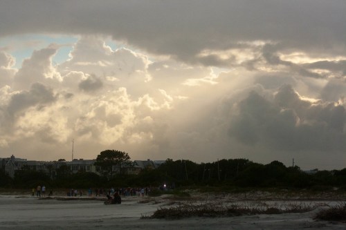 A beach scene with a crowd in the distance, under a dramatic sky filled with clouds and rays of sunlight.