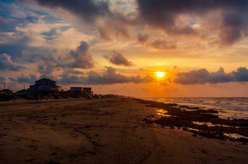 Sunset over a beach with clouds, waves, and coastal houses lining the shore.