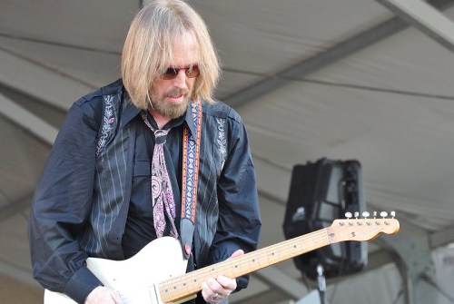 A musician with long hair and sunglasses plays an electric guitar on stage, wearing a black shirt and patterned tie.
