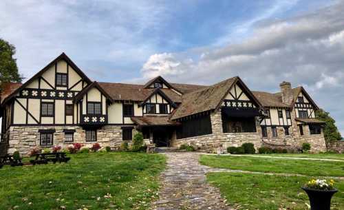 A large, historic Tudor-style house with stone and wood features, surrounded by green grass and a cloudy sky.