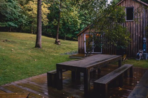 A wooden deck with a large table and benches, surrounded by trees and a rustic cabin in a serene outdoor setting.