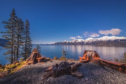 Scenic view of a calm lake surrounded by mountains, with wooden benches and trees in the foreground under a clear blue sky.