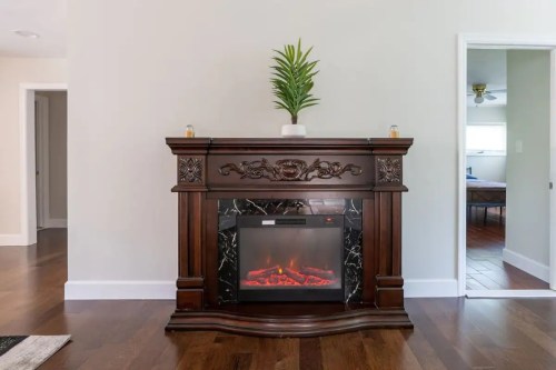 A decorative wooden fireplace with an electric insert and a potted plant on top, set against a light-colored wall.