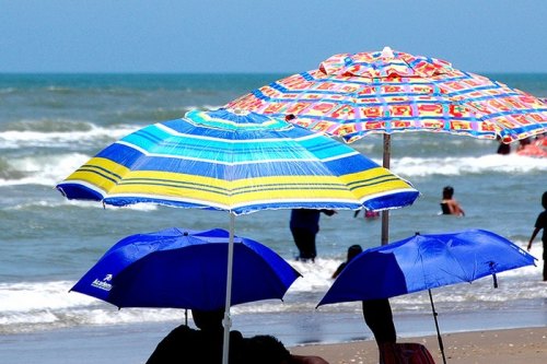 Colorful beach umbrellas shade people on a sandy shore, with waves and a clear blue sky in the background.