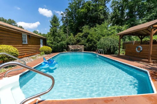 A clear swimming pool surrounded by greenery, with a blue inflatable toy and a wooden gazebo nearby.