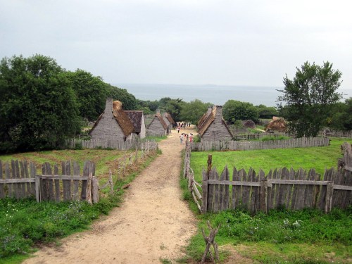 A scenic view of a historic village with thatched-roof houses, surrounded by greenery and a path leading to the ocean.