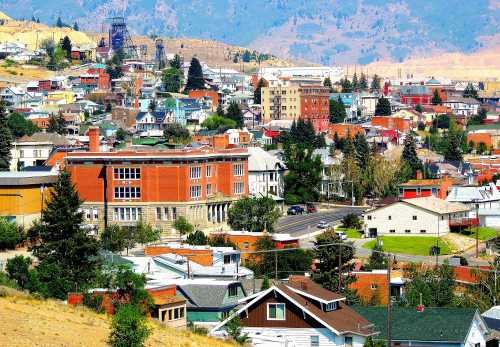 A colorful hillside town with various buildings, trees, and mountains in the background under a clear blue sky.