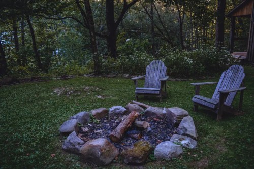 Two wooden chairs beside a stone fire pit with logs, surrounded by trees in a serene outdoor setting.
