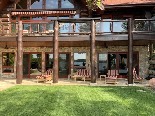 A rustic log cabin with large windows, stone walls, and striped lounge chairs on a grassy patio overlooking a lake.