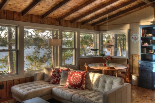 Cozy cabin interior with a white sofa, wooden beams, and a dining table by large windows overlooking a lake.