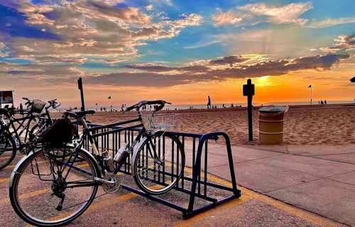 A bicycle parked at a beachside rack with a vibrant sunset and clouds in the background.