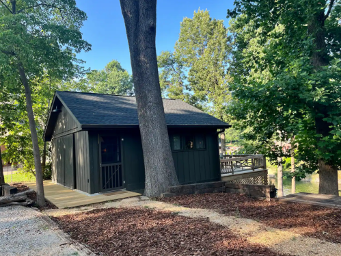 A modern dark green cabin surrounded by trees, with a wooden deck and gravel pathway leading to it.