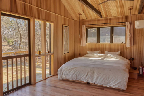 Cozy wooden bedroom with a large bed, windows, and a view of nature outside. Warm, inviting atmosphere.