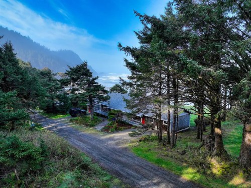A coastal house surrounded by trees, with a view of the ocean and mountains in the background under a clear blue sky.