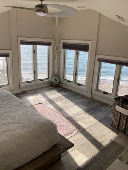Bright bedroom with a bed, large windows overlooking the beach, and sunlight casting shadows on the floor.