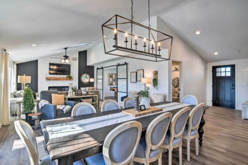 A modern dining area with a long table, stylish chairs, and a large chandelier, leading to a cozy living space.