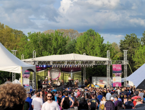 A lively outdoor concert scene with a crowd enjoying a performance on stage, surrounded by trees and tents.