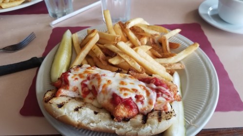 A plate featuring a breaded chicken sandwich topped with marinara and cheese, served with fries and pickles.