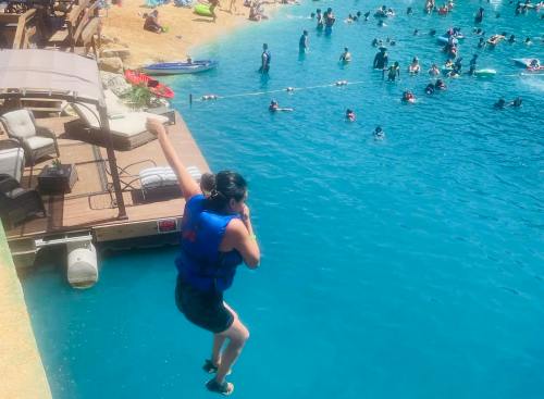 A person in a life jacket jumps off a dock into a blue pool filled with swimmers and floating devices.