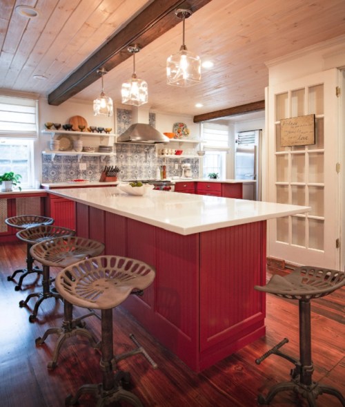 A modern kitchen with red cabinets, a white island, pendant lights, and vintage-style stools.