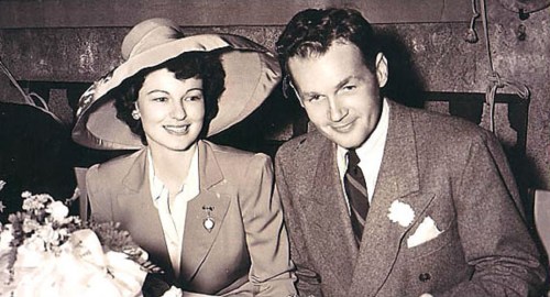 A smiling couple seated together, the woman wearing a large hat and the man in a suit, with flowers in front of them.
