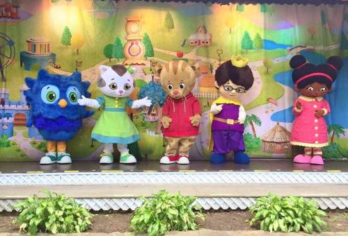 Five colorful mascots stand on stage, including an owl, a cat, a bear, a boy in glasses, and a girl in a pink dress.