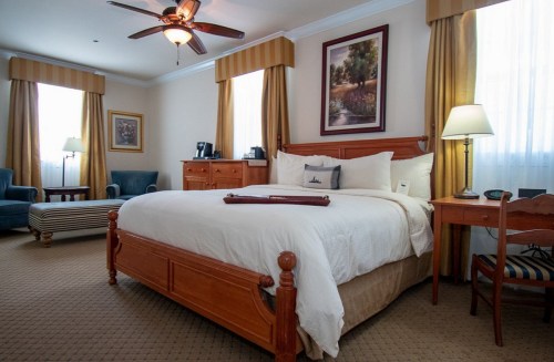 Cozy hotel room featuring a wooden bed, armchairs, desk, and large windows with curtains. Warm, inviting decor.