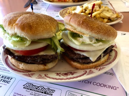 Two stacked hamburgers with lettuce, tomato, and cheese on a plate, accompanied by fries in the background.