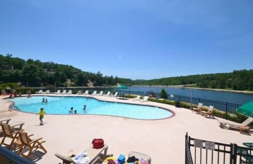 A sunny pool area with people swimming, surrounded by lounge chairs and a scenic view of a lake and greenery.