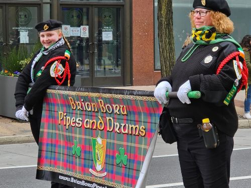 Two women in black uniforms hold a colorful banner for "Briann Boru Pipes and Drums" at a parade.