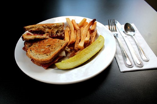 A plate with a toasted sandwich, fries, and a pickle, set on a black table with utensils beside it.