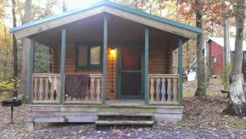 A cozy wooden cabin with a porch, surrounded by autumn trees and a gravel path. Warm light glows from the entrance.
